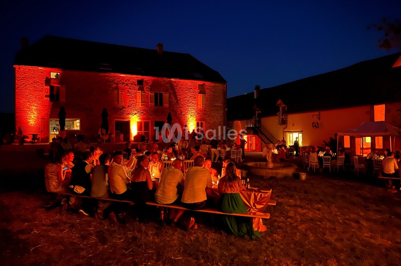 Groupe de personnes dînant en plein air devant un bâtiment en pierre éclairé par des lumières rouges, de nuit.