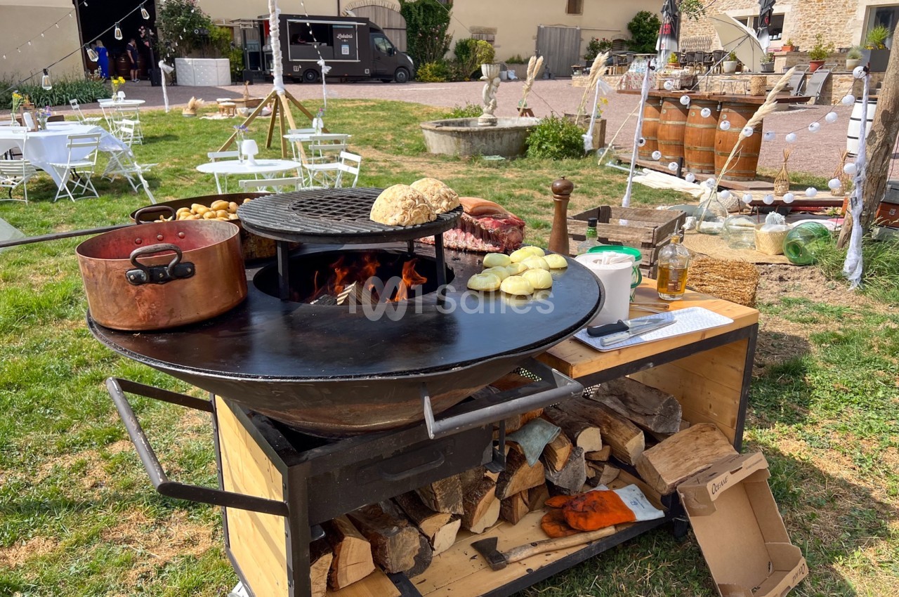 Barbecue circulaire en plein air avec légumes, pain et marmite en cuivre, entouré d'une ambiance festive champêtre.