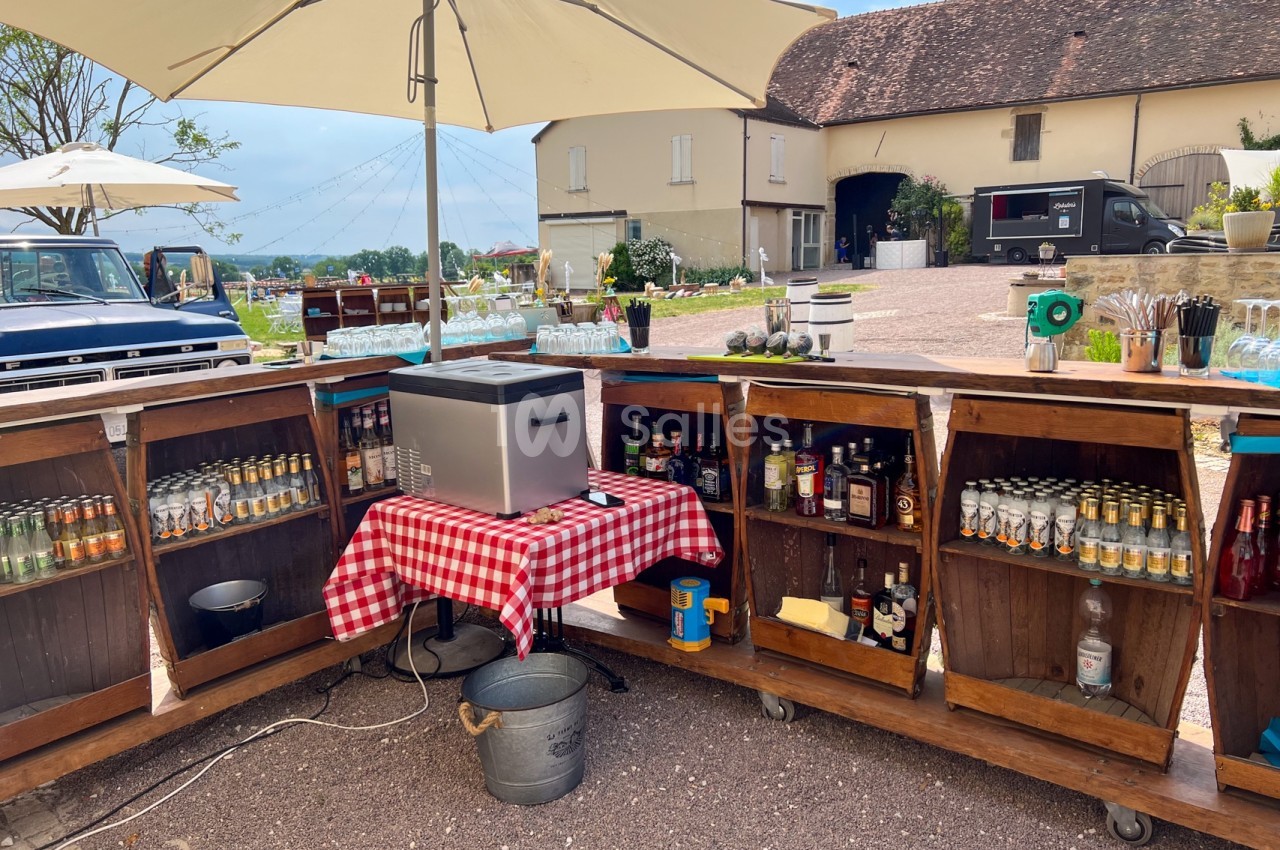 Bar extérieur en bois avec boissons et verres, installé sous un parasol dans une cour pavée devant des bâtiments anciens.