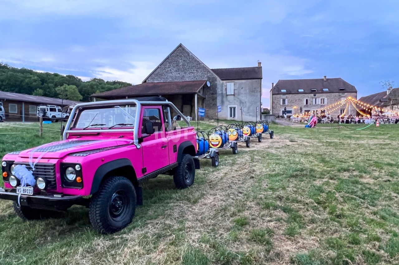 Un 4x4 rose tracte un petit train décoré dans un champ près de bâtiments en pierre, avec une fête éclairée en arrière-plan.