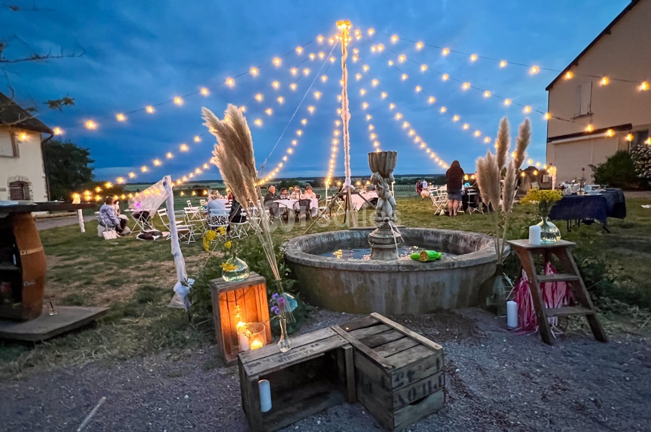Cour extérieure décorée avec une fontaine centrale, guirlandes lumineuses et tables dressées pour un événement en soirée.