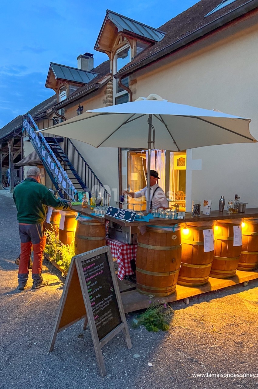 Stand extérieur éclairé en soirée, avec bar en tonneaux, parasol blanc et ardoise de menu devant une maison en pierre.