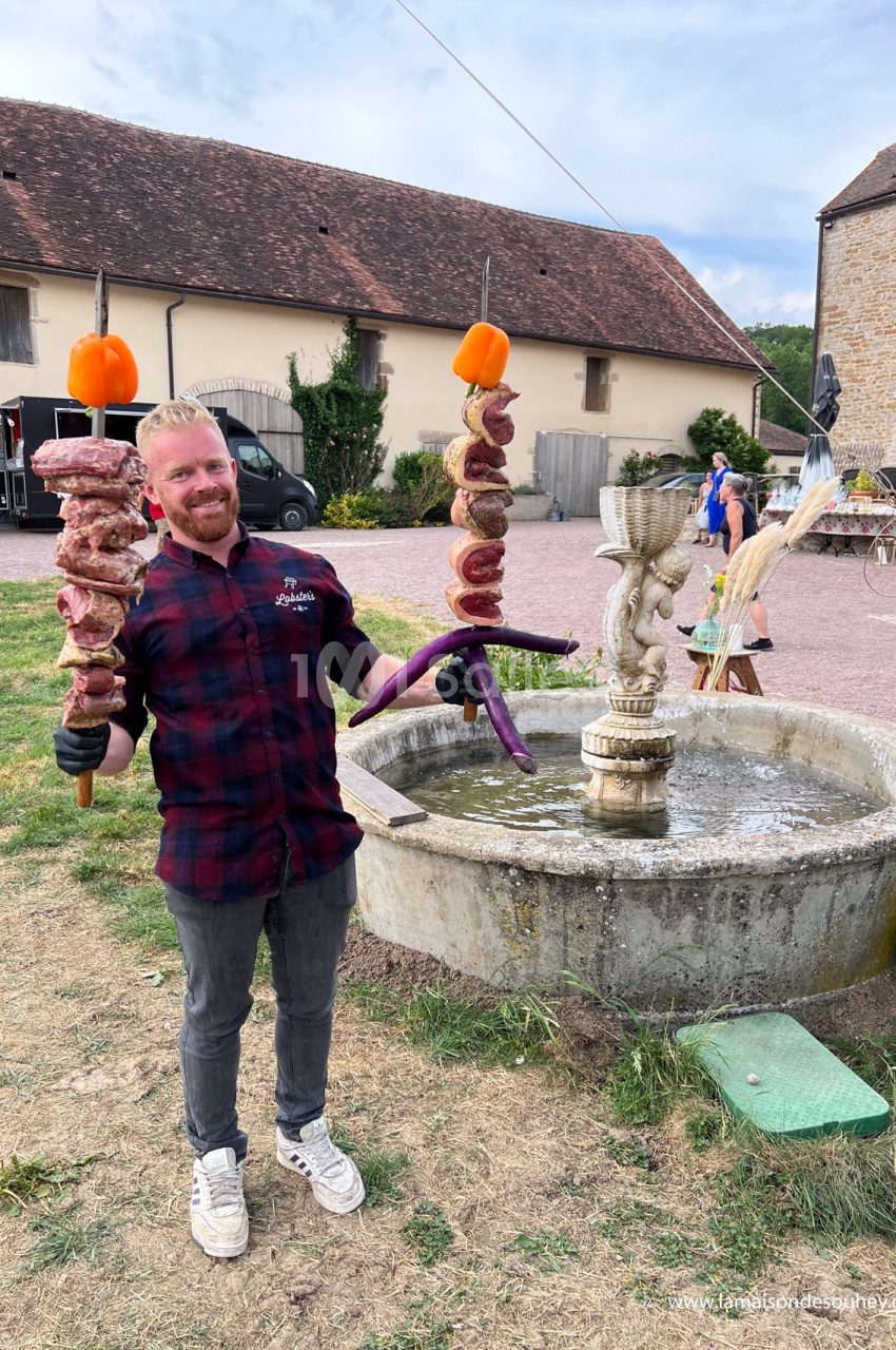 Homme souriant tenant des brochettes géantes devant une fontaine dans un cadre rural.