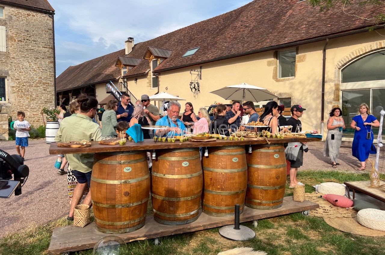 Des invités se rassemblent autour d'un buffet disposé sur des tonneaux en extérieur devant un bâtiment en pierre.