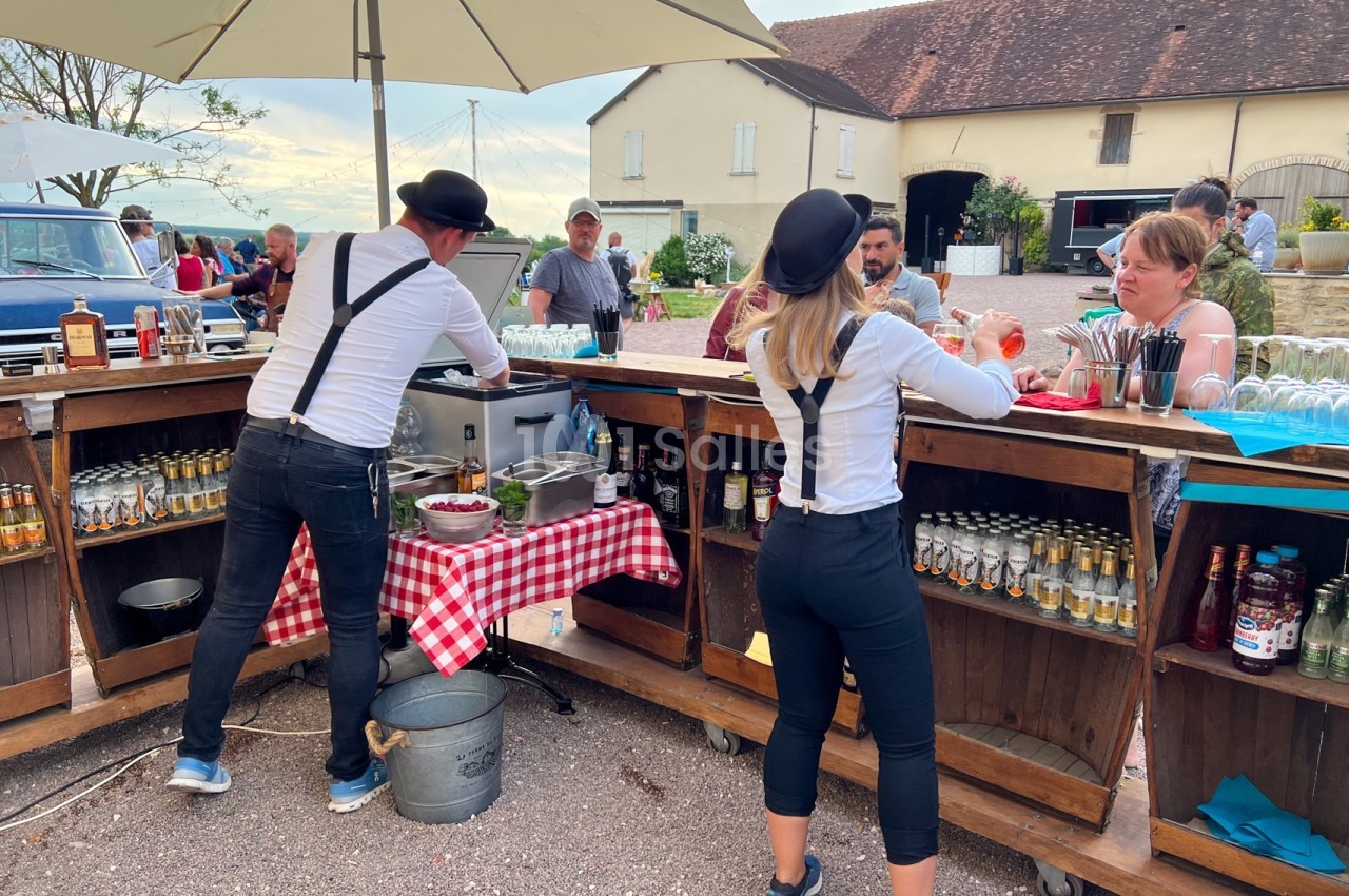 Deux personnes en tenue rétro servent des boissons derrière un bar en bois lors d'un événement en plein air.