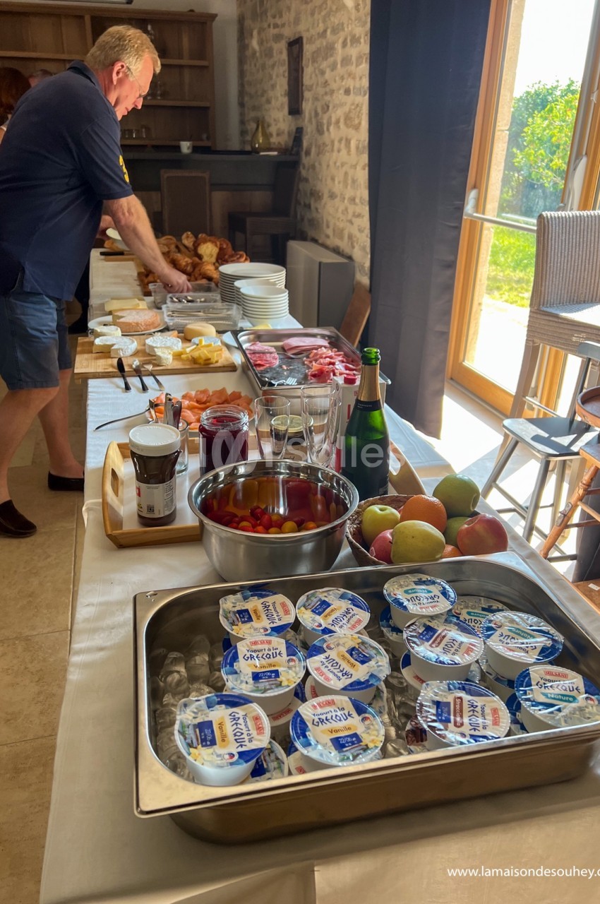Un buffet de petit-déjeuner avec yaourts, fruits, viennoiseries, charcuterie et boissons, dans une salle lumineuse.