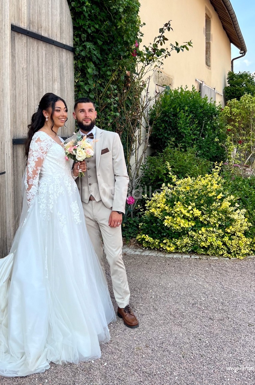 Un couple en tenue de mariage pose devant un bâtiment en pierre entouré de végétation.