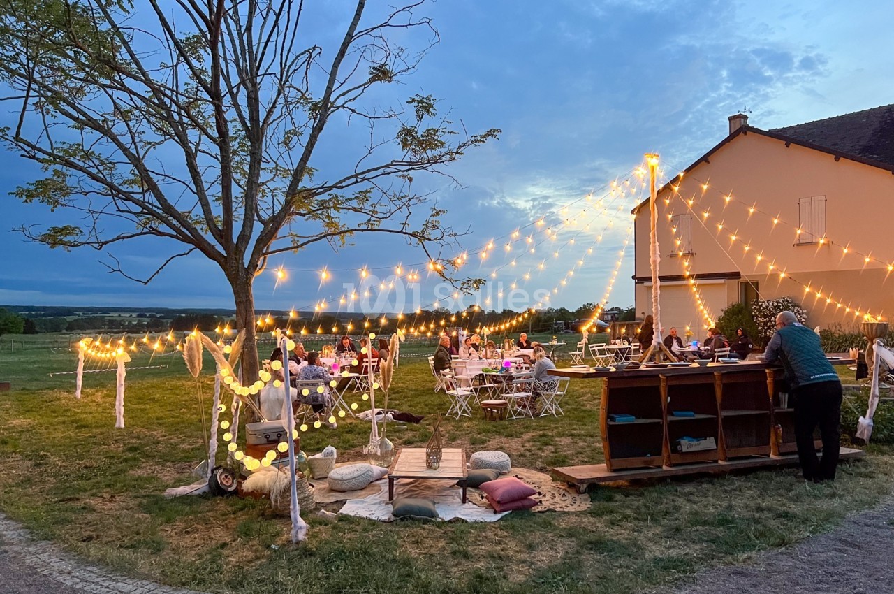 Espace extérieur décoré avec guirlandes lumineuses, tables dressées et bar en bois devant une maison au crépuscule.