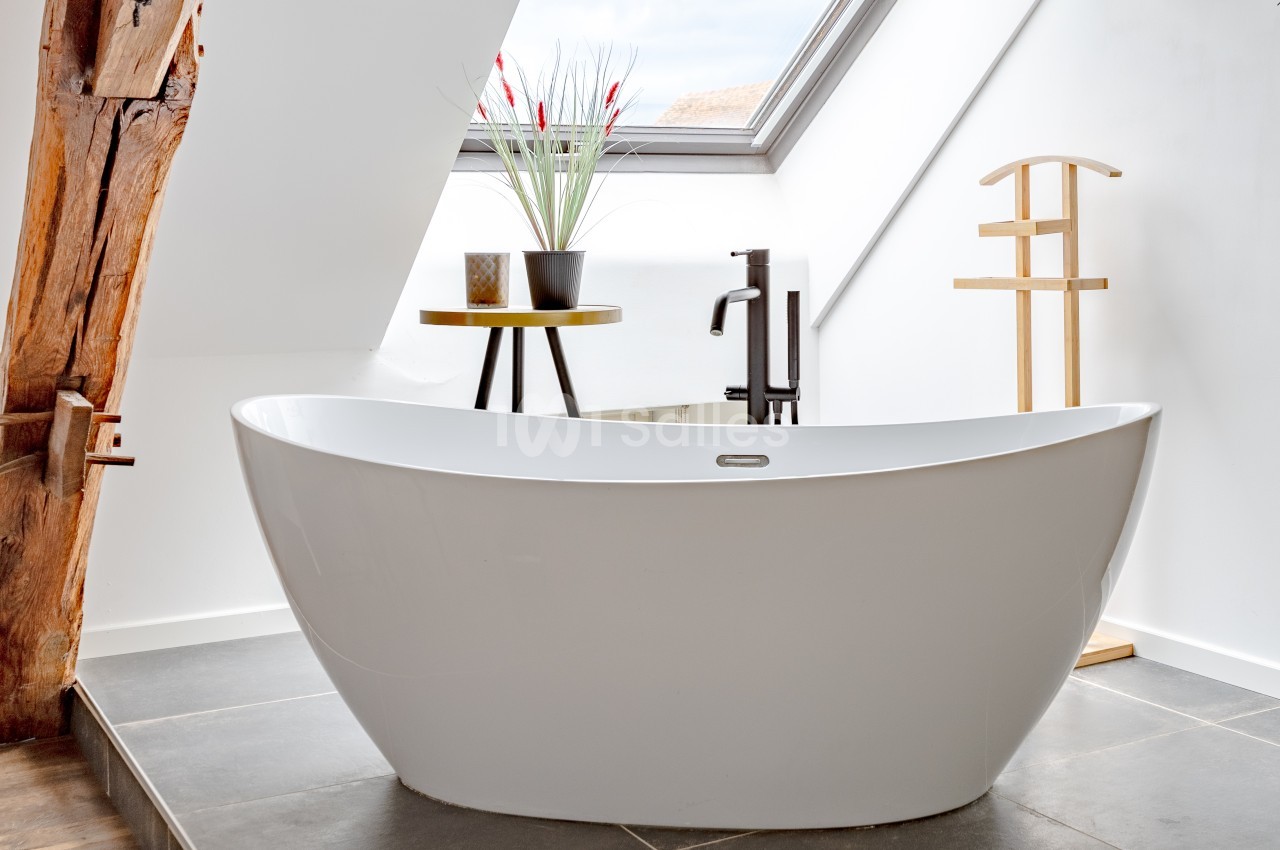 Baignoire blanche moderne dans une salle de bain lumineuse avec fenêtre de toit, plantes et porte-serviettes en bois.