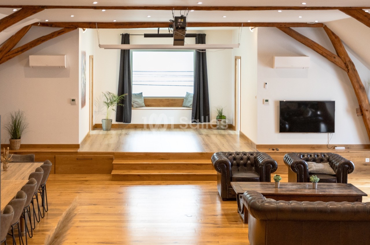 Salle lumineuse avec parquet, canapés en cuir, table en bois, et espace surélevé avec fenêtre et rideaux noirs.