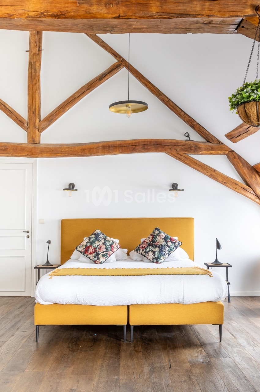 Lit double avec tête de lit jaune moutarde, poutres apparentes en bois, suspension et décoration murale dans une chambre…