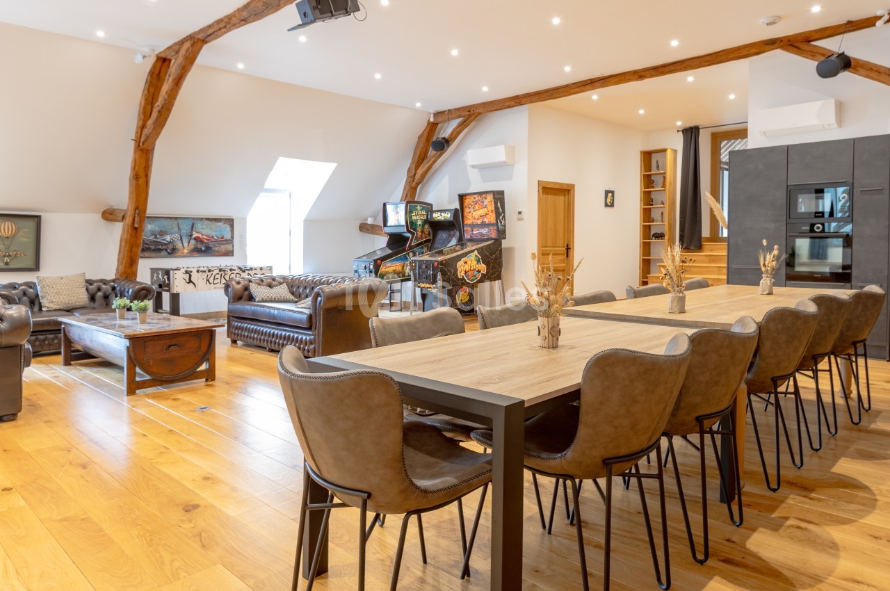 Salle lumineuse avec table en bois, chaises en cuir, canapés Chesterfield, flippers et poutres apparentes.