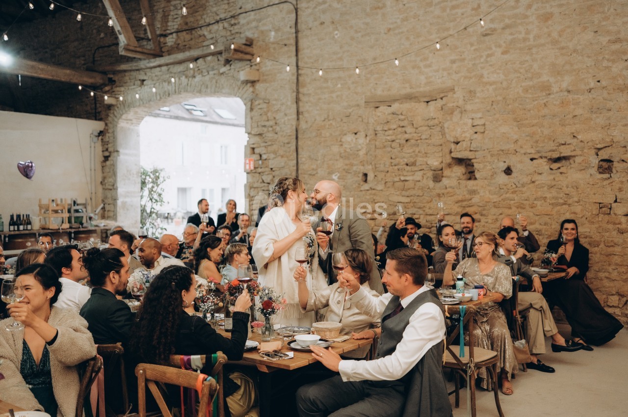Un couple échange un baiser lors d'un repas de mariage dans une salle en pierre, entouré d'invités souriants.