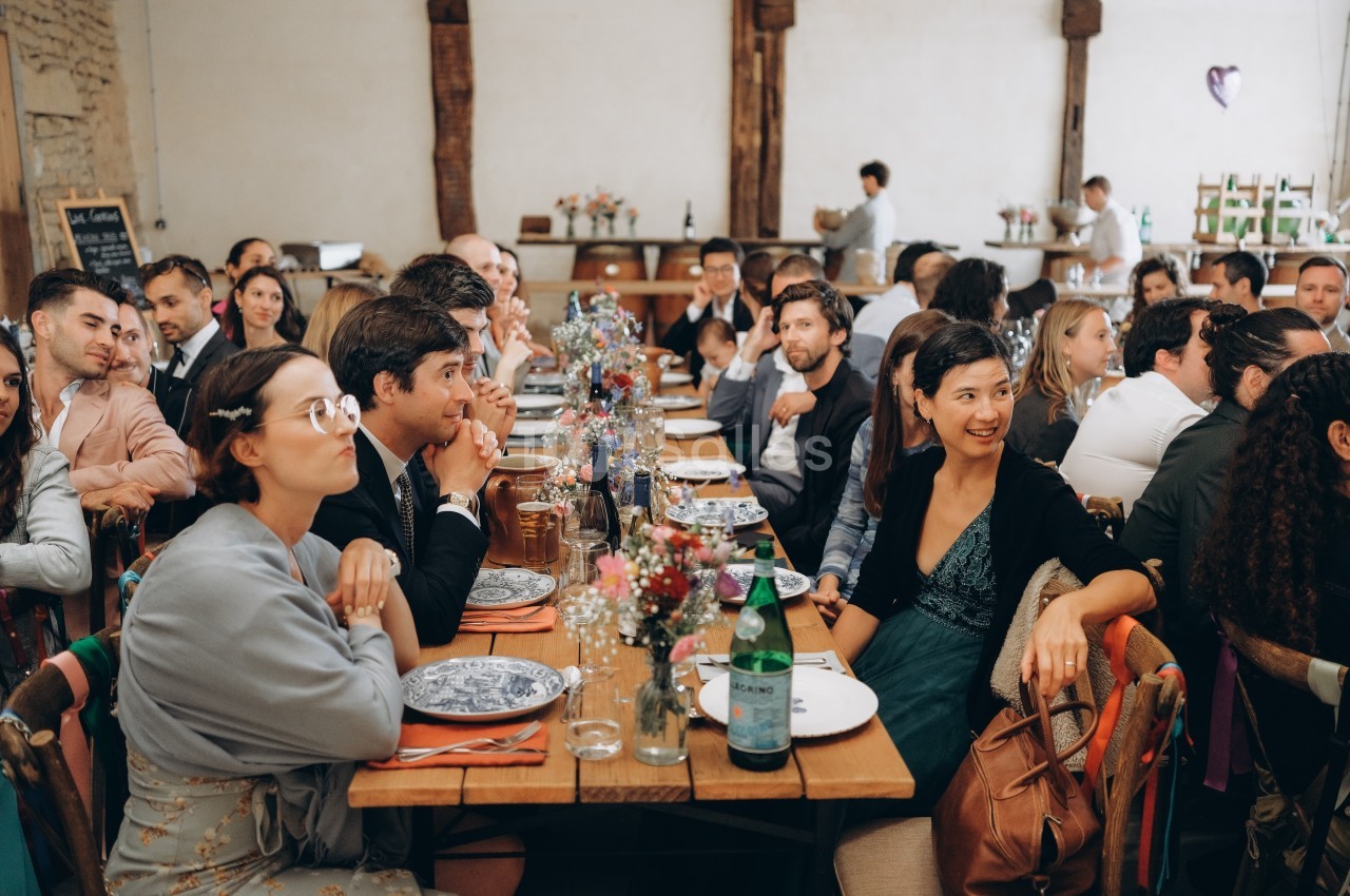 Des convives assis à une longue table décorée, lors d'un repas dans une salle au style rustique.
