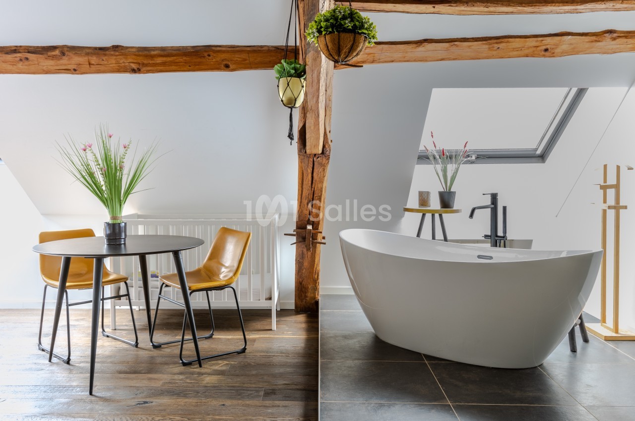 Salle de bain lumineuse avec baignoire îlot, table ronde, chaises en cuir et poutres apparentes sous un toit mansardé.