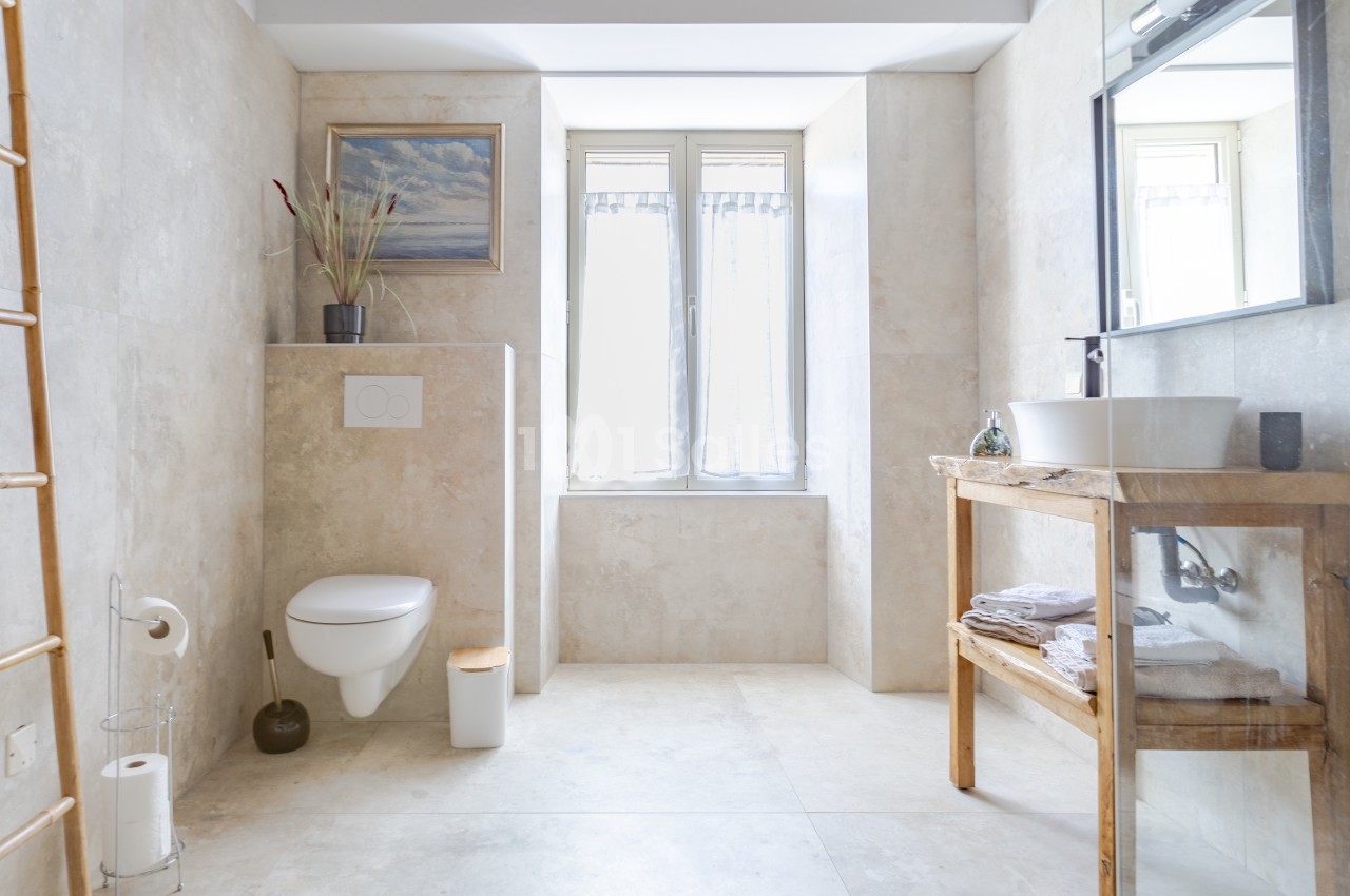 Salle de bain lumineuse avec toilettes suspendues, vasque sur meuble en bois et fenêtre laissant entrer la lumière naturelle.