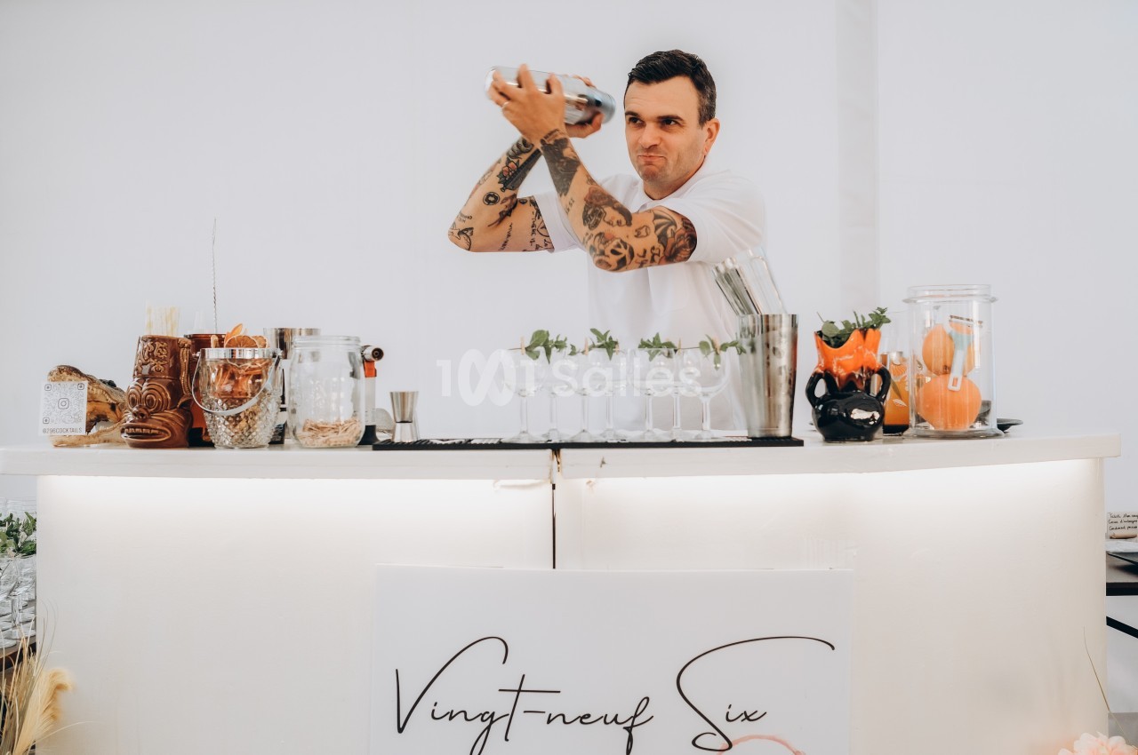 Un barman tatoué prépare un cocktail en secouant un shaker derrière un comptoir blanc décoré de verres et d'ingrédients.