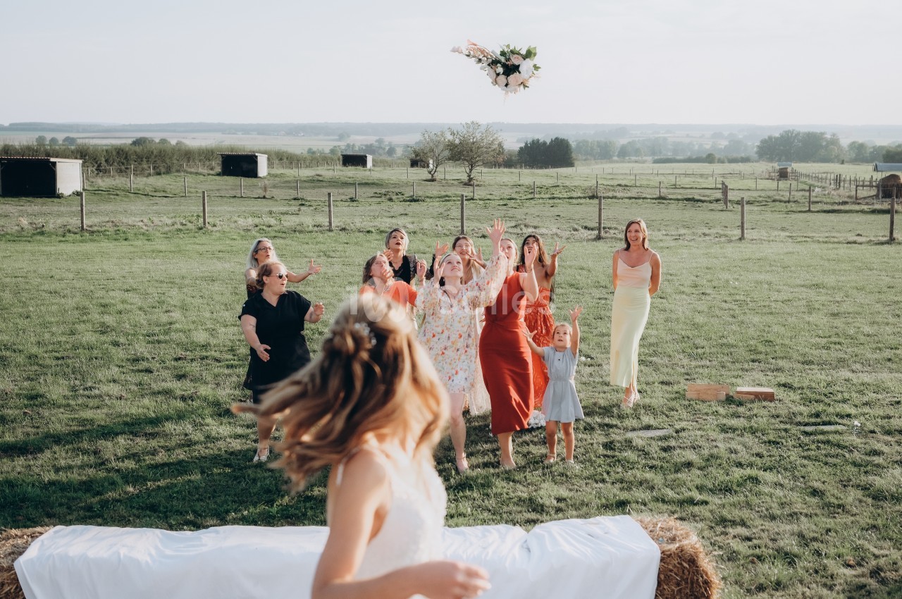 Une mariée lance son bouquet à un groupe de femmes dans un champ lors d'une réception en plein air.