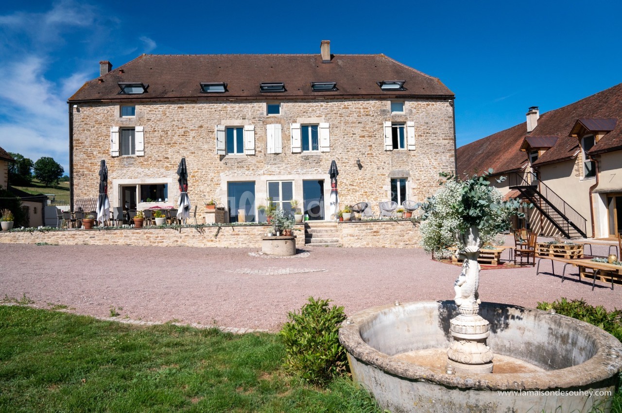 Façade d'une grande maison en pierre avec terrasse, parasols et une fontaine au premier plan, sous un ciel bleu.