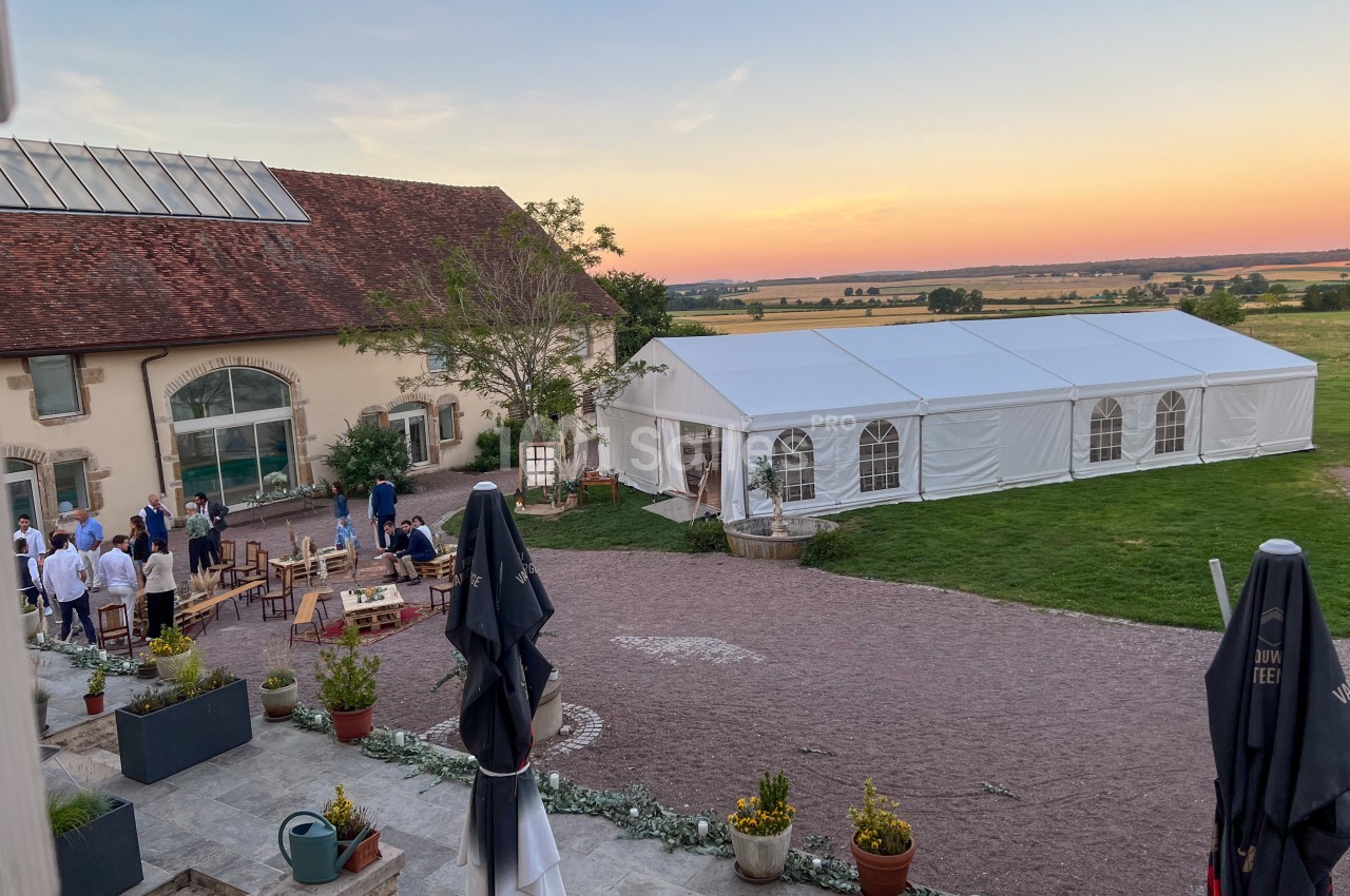 Cour d'une propriété avec une tente blanche, des invités rassemblés et un paysage champêtre au coucher du soleil.