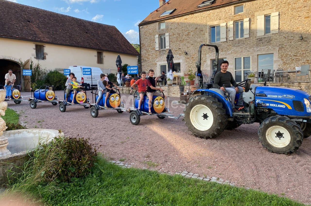 Un tracteur bleu tire un petit train de barils décorés avec des visages souriants, devant des bâtiments en pierre.