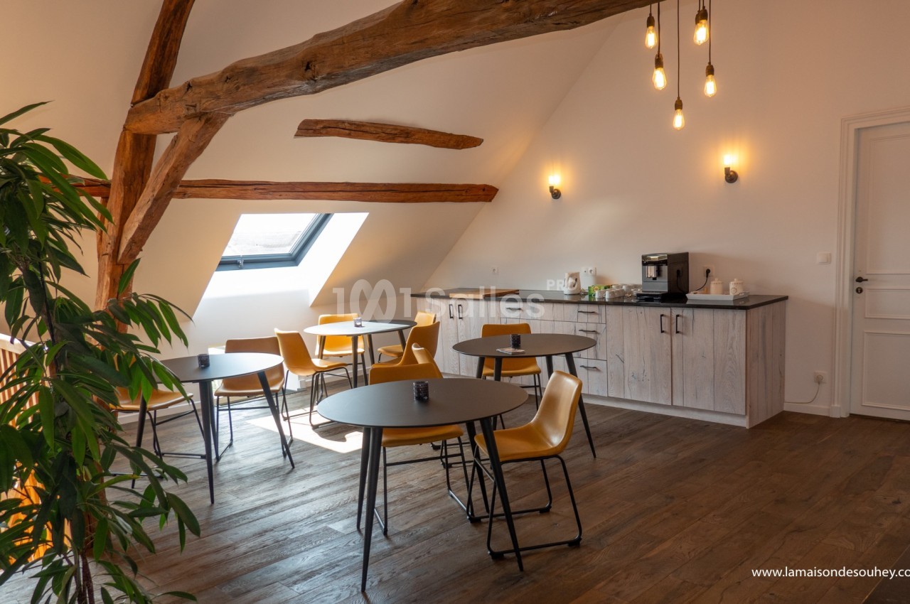Salle lumineuse sous combles avec tables, chaises jaunes, poutres apparentes, kitchenette et plantes décoratives.