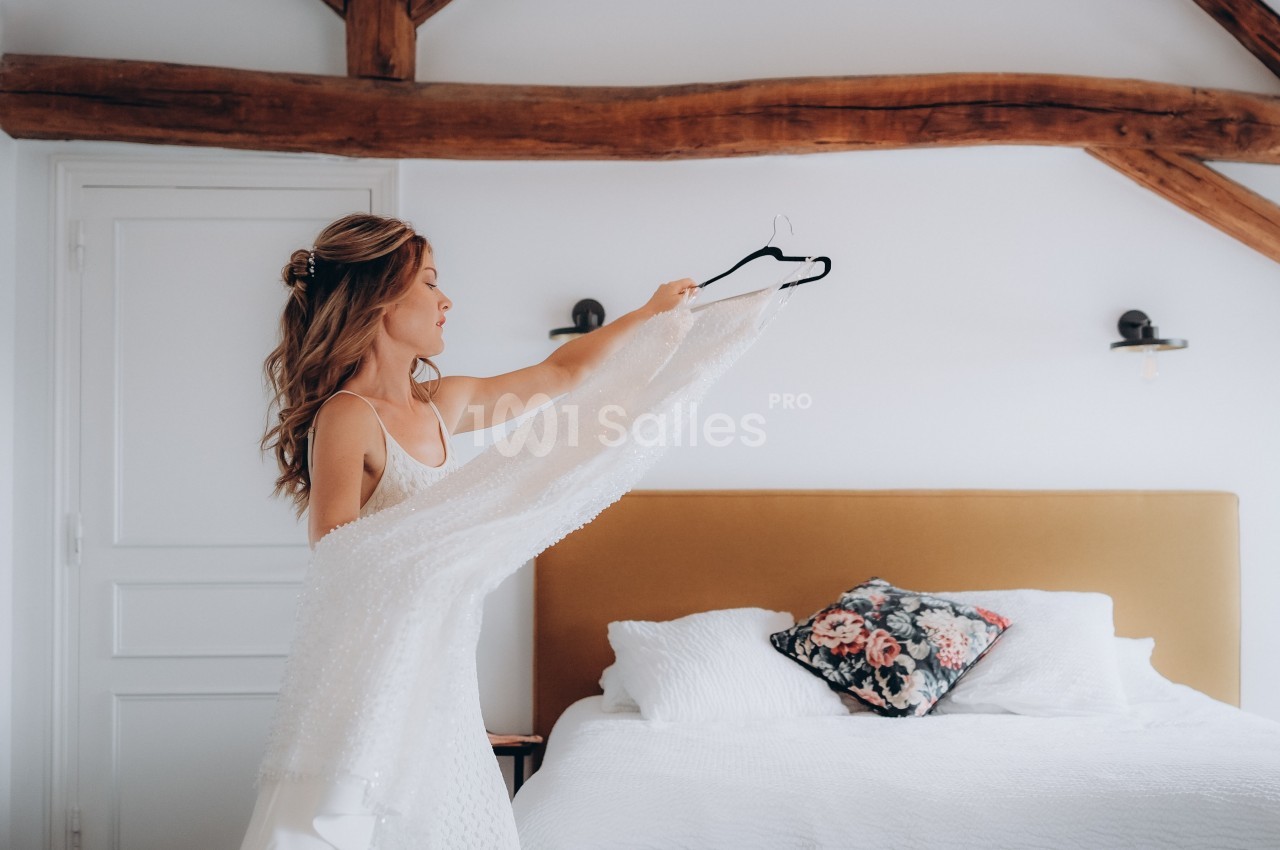 Une femme tient une robe blanche sur un cintre dans une chambre lumineuse avec un lit et des poutres apparentes.