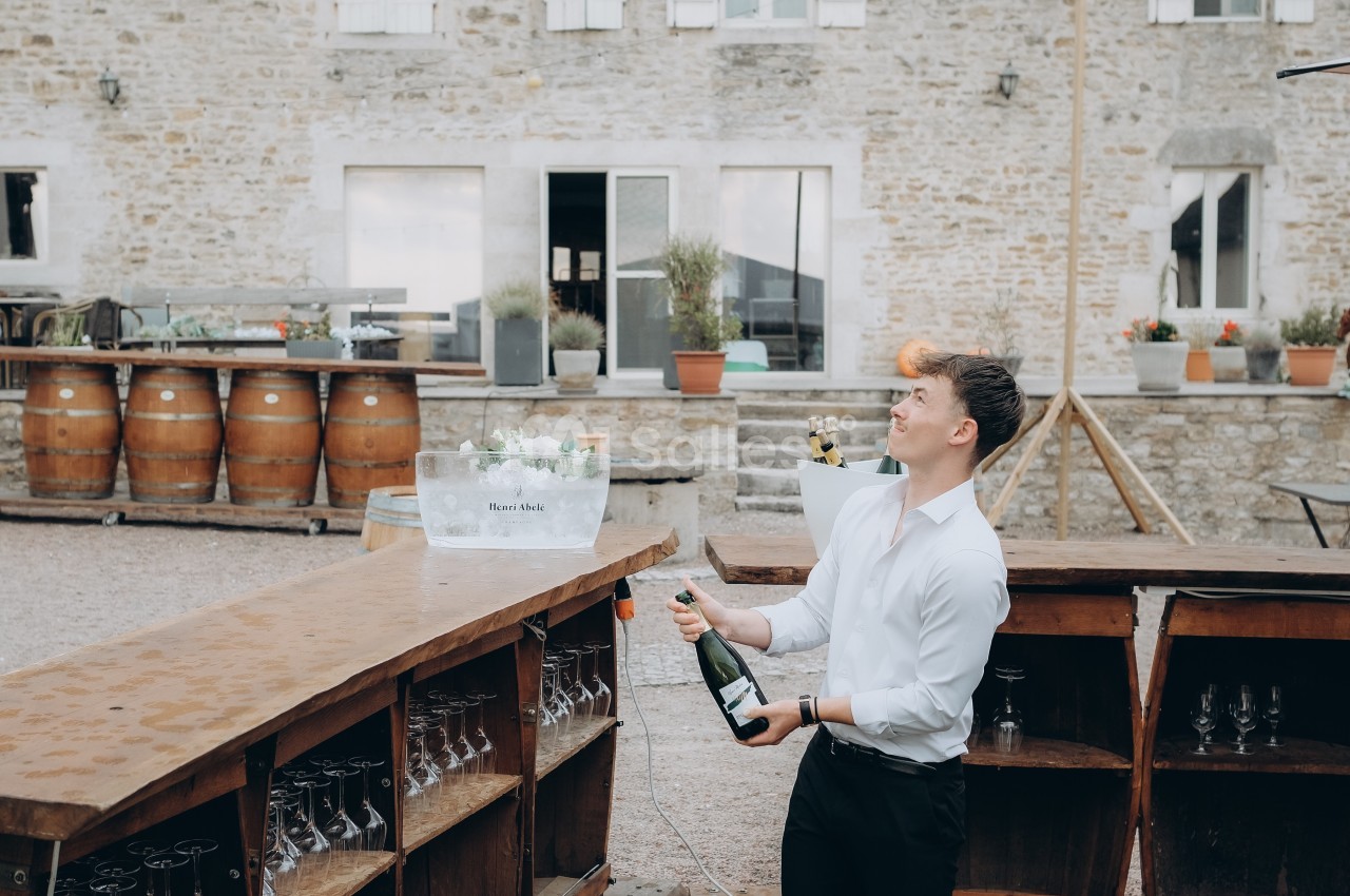 Un homme ouvre une bouteille de champagne derrière un bar extérieur, devant un bâtiment en pierre.