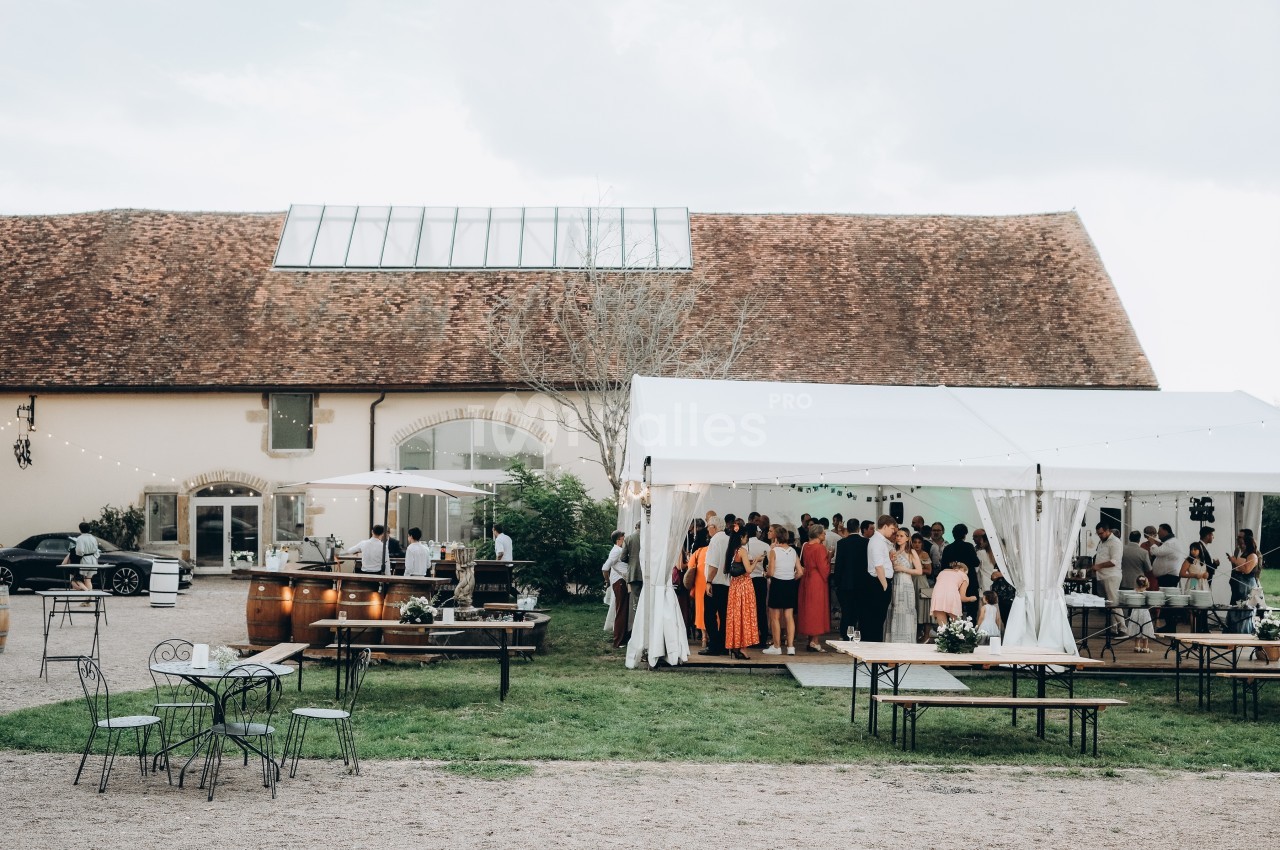 Groupe de personnes rassemblées sous une tente blanche dans une cour avec tables, chaises et bâtiment en arrière-plan.