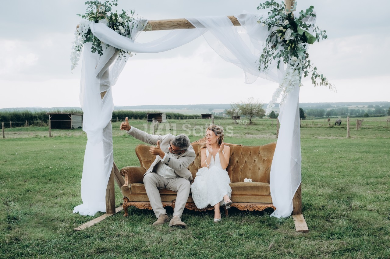 Un couple assis sur un canapé en plein air sous une arche décorée de voilages et de fleurs, dans un champ.