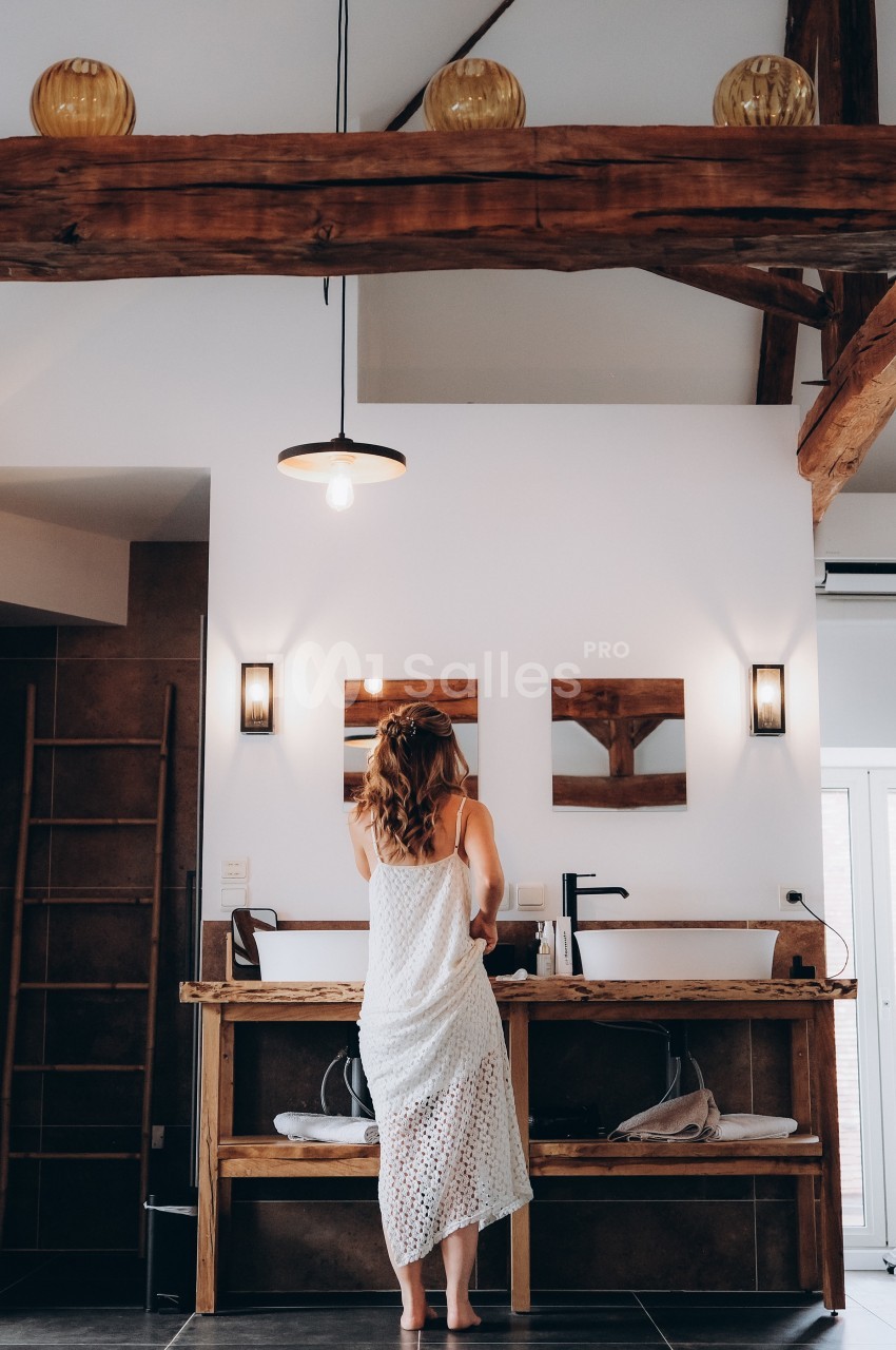 Femme de dos en robe blanche se tenant devant un lavabo dans une salle de bain au style rustique avec poutres apparentes.
