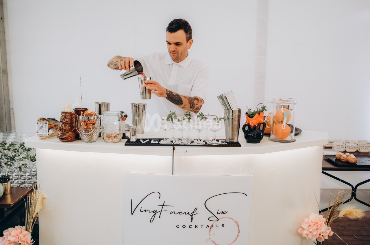 Un barman prépare un cocktail derrière un comptoir blanc décoré avec des ingrédients et des accessoires.