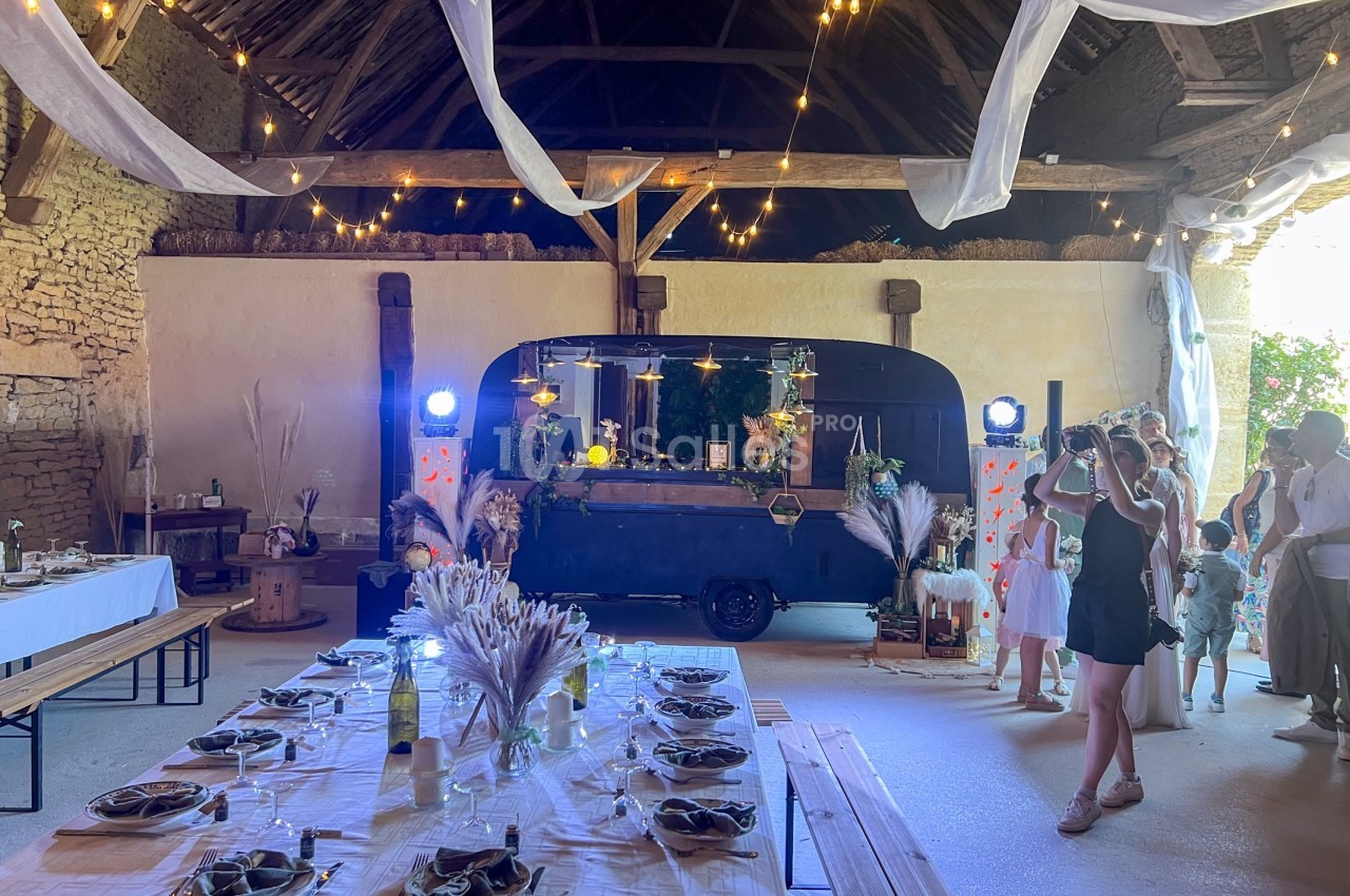 Salle de réception décorée pour un événement, avec tables dressées, guirlandes lumineuses et un bar mobile en arrière-plan.