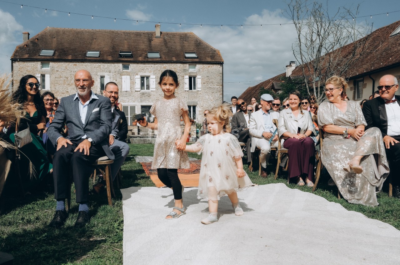 Deux enfants marchent main dans la main sur une allée blanche lors d'une cérémonie en extérieur, entourés d'invités…