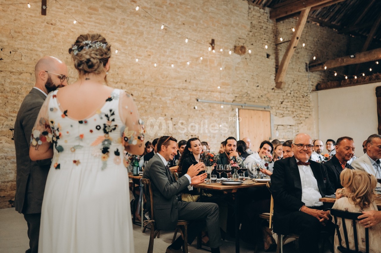 Un couple en tenue de mariage s'adresse à des invités assis dans une salle en pierre décorée de guirlandes lumineuses.