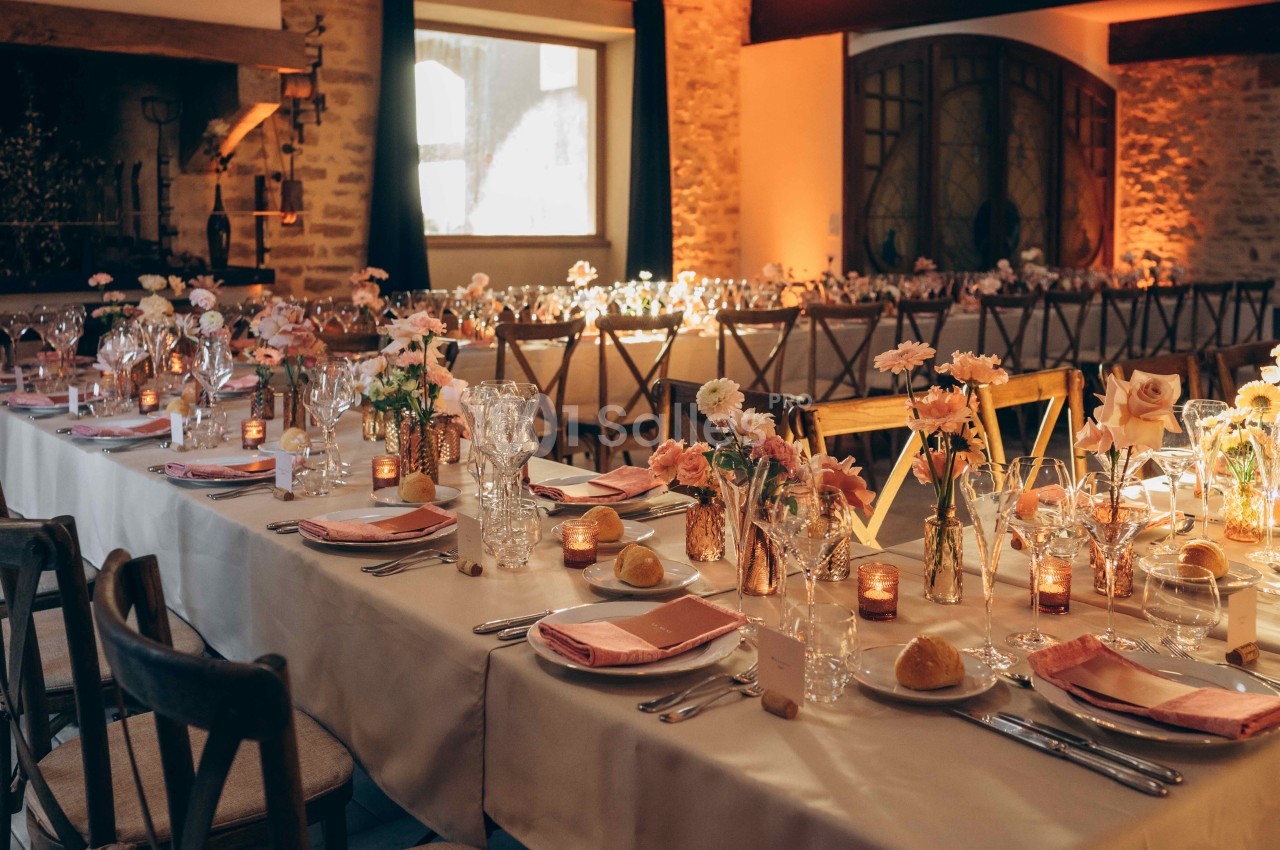 Salle de réception décorée avec des tables élégantes, nappes blanches, fleurs pastel et bougies allumées.