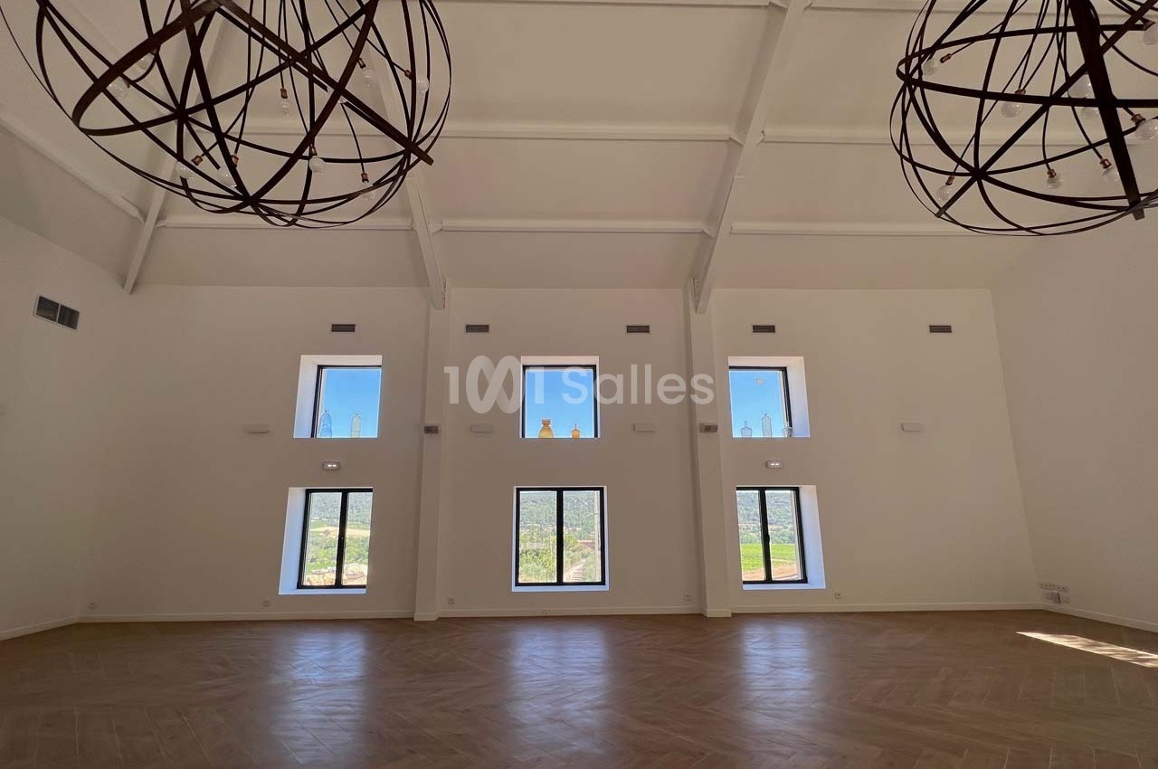 Grande salle lumineuse avec parquet en bois clair, hauts plafonds blancs et grandes fenêtres offrant une vue sur l'extérieur.