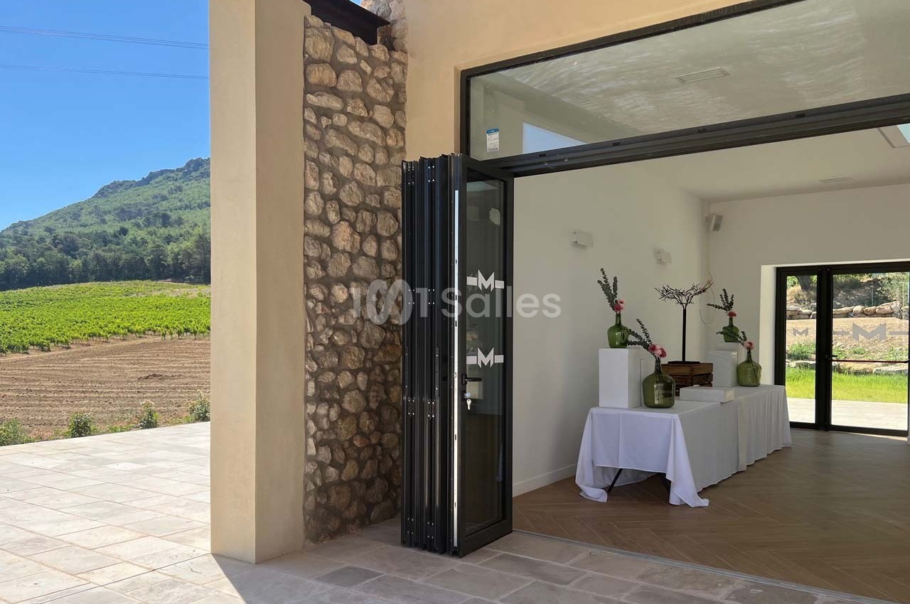 Entrée d'une salle lumineuse avec table décorée, ouverte sur une terrasse en pierre et un paysage de vignobles.