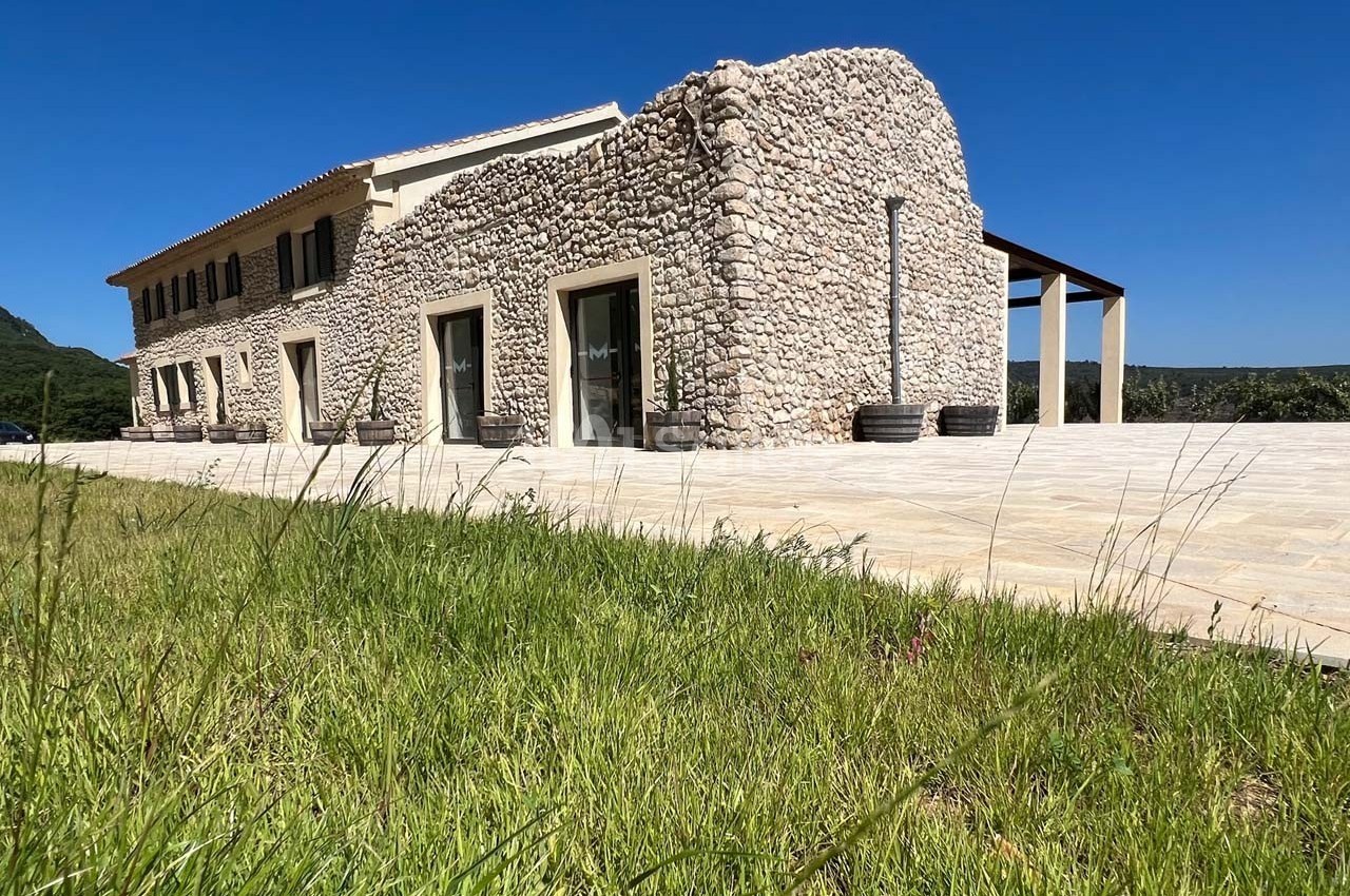 Façade d'une maison en pierre avec grandes baies vitrées, terrasse pavée et pelouse au premier plan.
