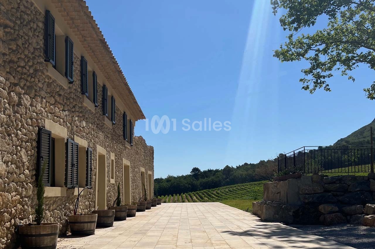 Façade en pierre d'une maison avec volets noirs, terrasse pavée et vignoble en arrière-plan sous un ciel bleu.