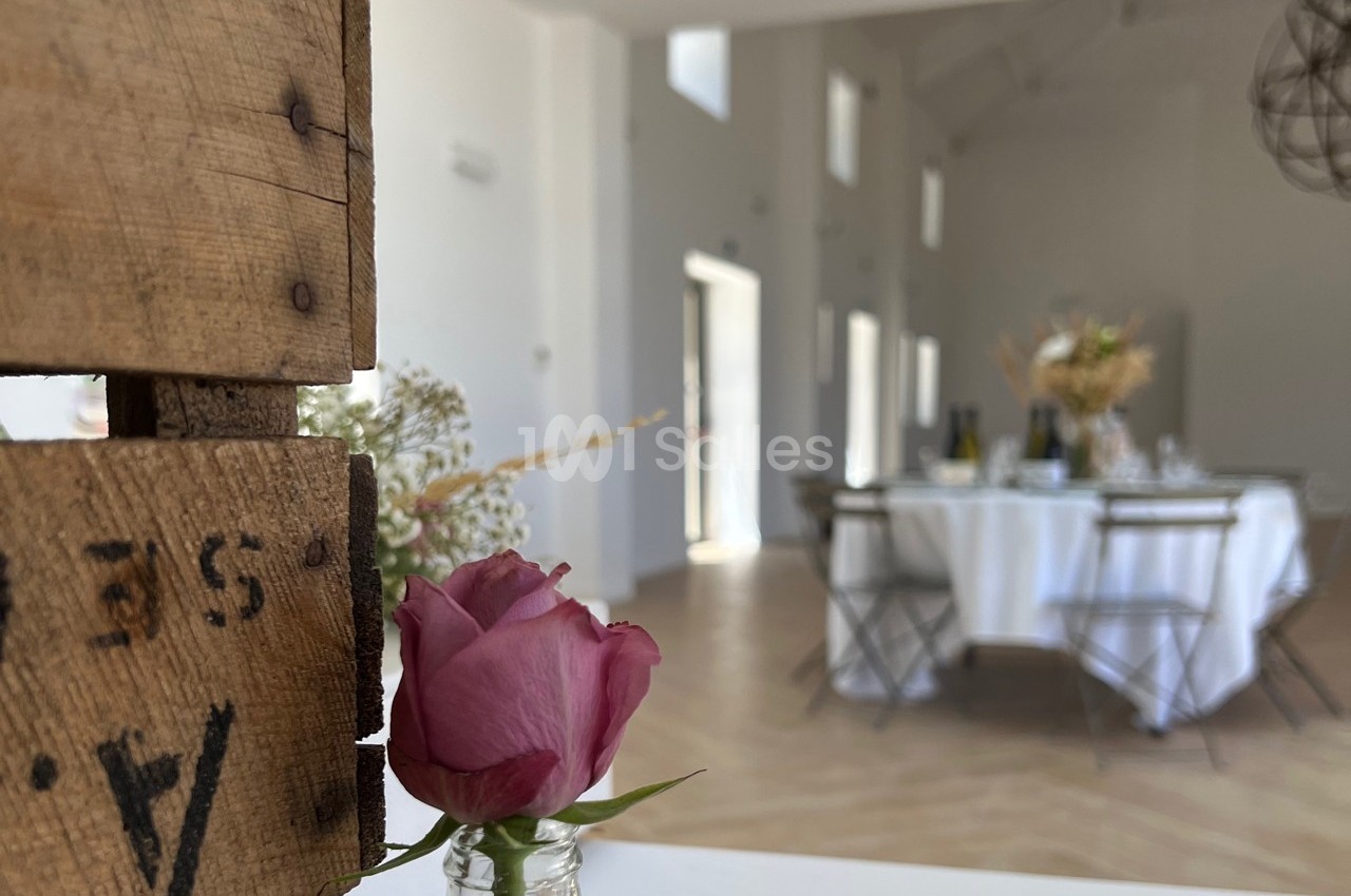 Rose rose en gros plan dans un vase, avec en arrière-plan une salle lumineuse aménagée pour un repas.