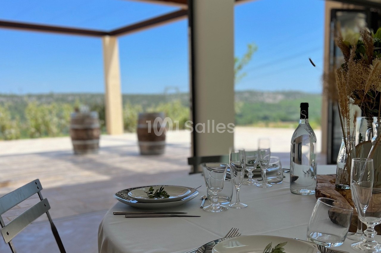 Table dressée avec vaisselle élégante, vue sur une terrasse ensoleillée et des barriques en arrière-plan.