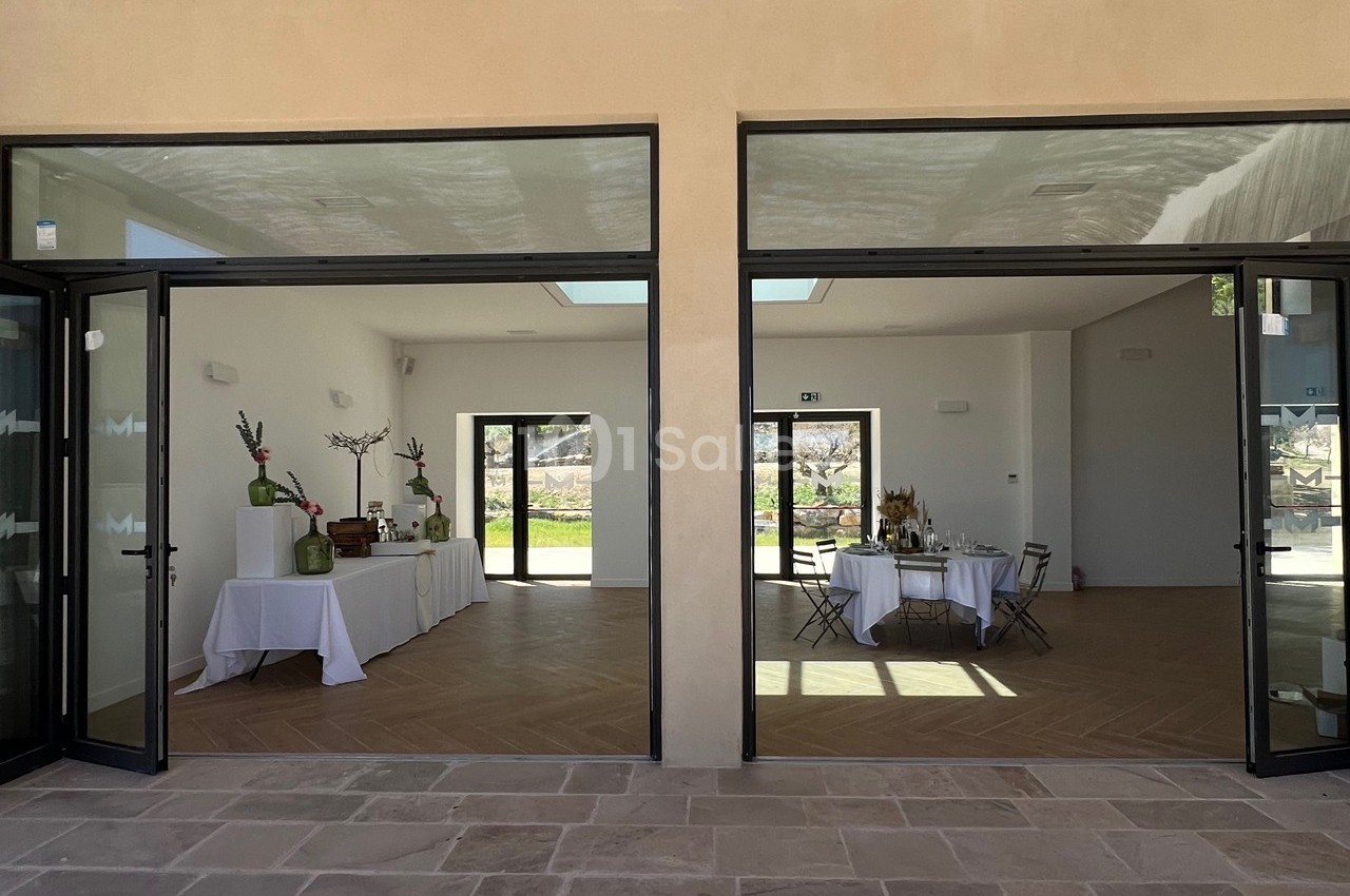 Salle lumineuse avec grandes baies vitrées, tables dressées et décorations minimalistes, vue sur un extérieur ensoleillé.