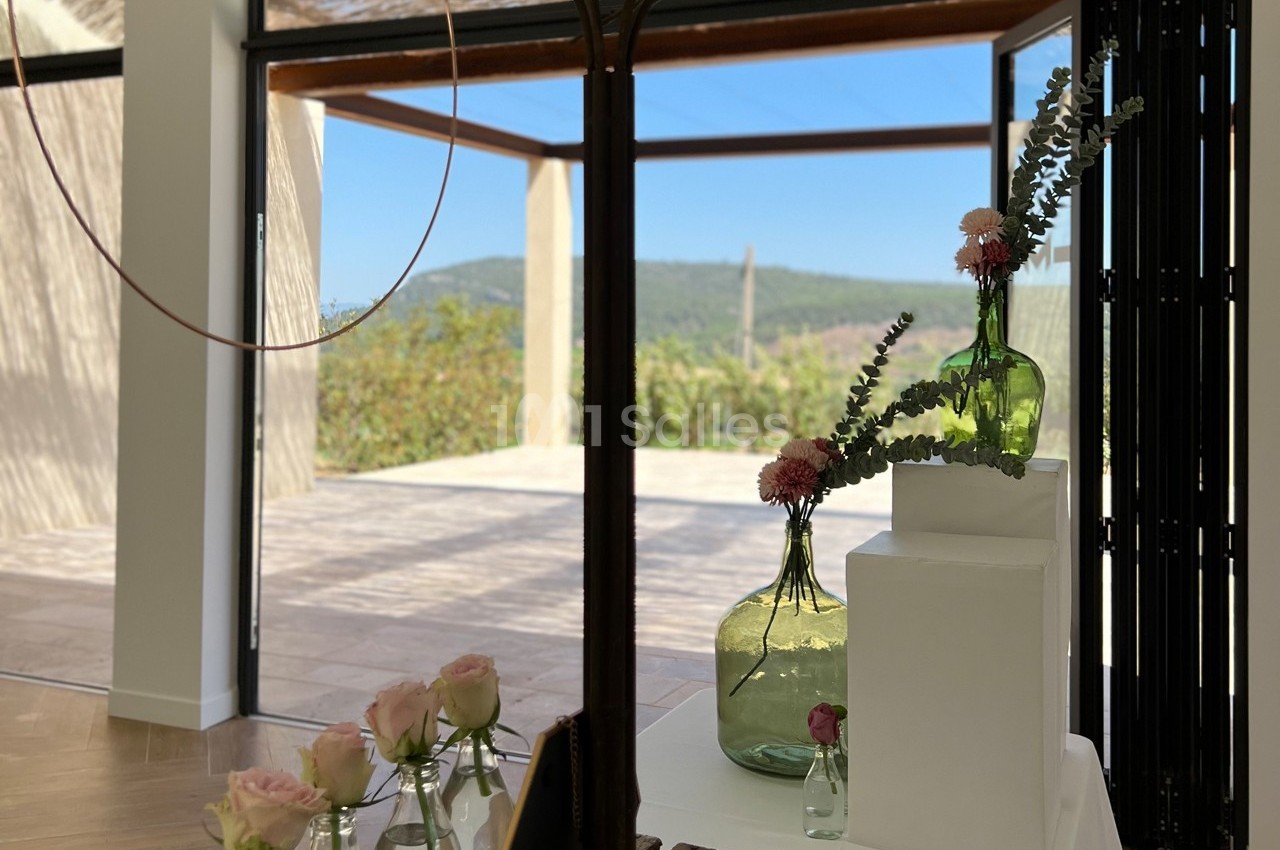 Vases en verre avec fleurs roses et eucalyptus, posés sur des supports blancs, devant une terrasse ensoleillée avec vue sur…