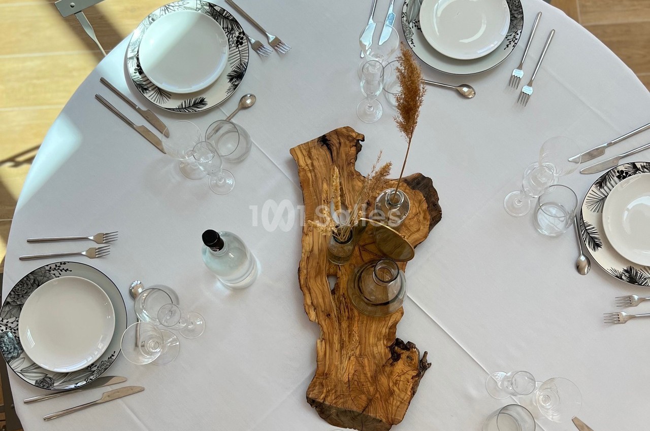 Table dressée avec vaisselle blanche et noire, couverts en métal, décoration centrale en bois et herbes sèches.