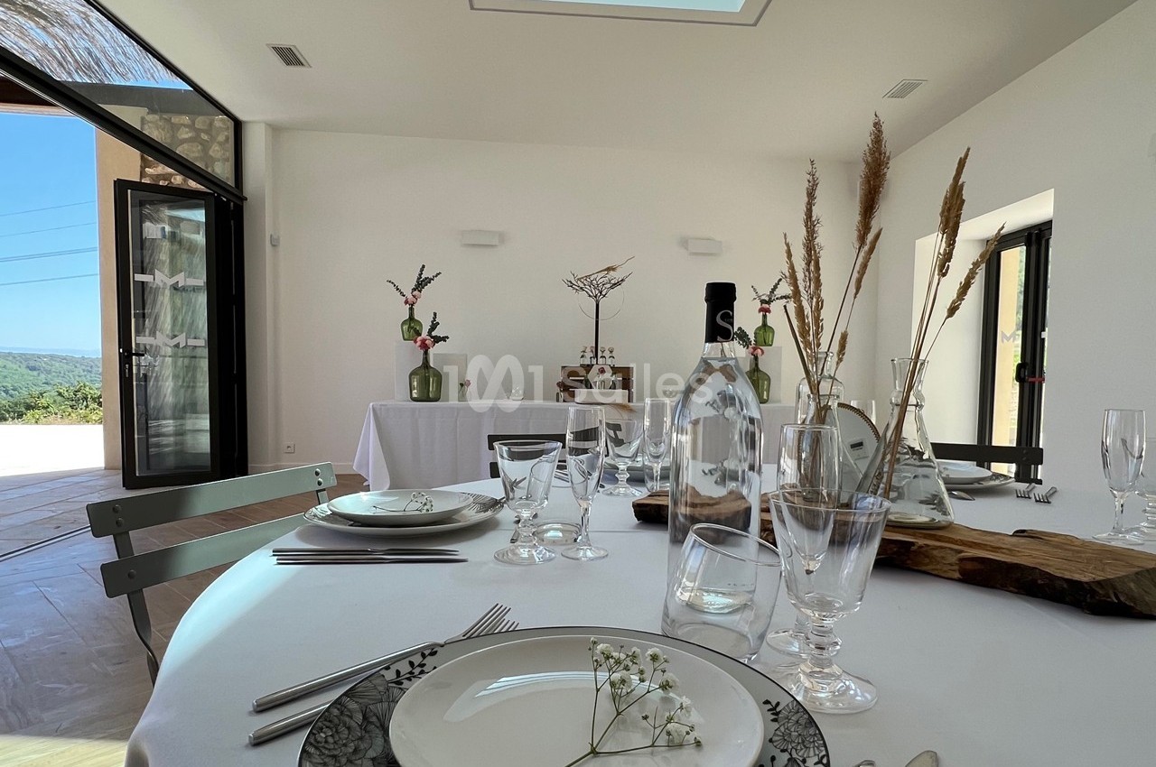 Table dressée avec vaisselle élégante dans une salle lumineuse, décorée de fleurs et ouverte sur un paysage extérieur.