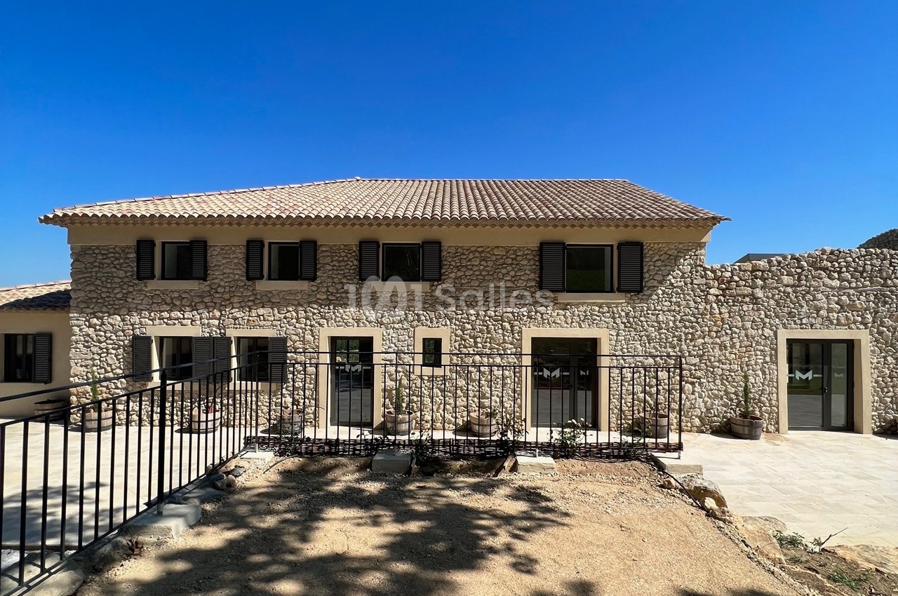 Façade d'une maison en pierre avec volets noirs, terrasse ensoleillée et sol en terre devant une clôture métallique.
