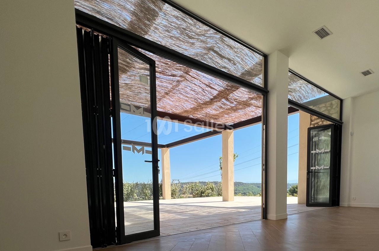 Baie vitrée ouverte sur une terrasse ombragée par une pergola, offrant une vue dégagée sur un paysage ensoleillé.