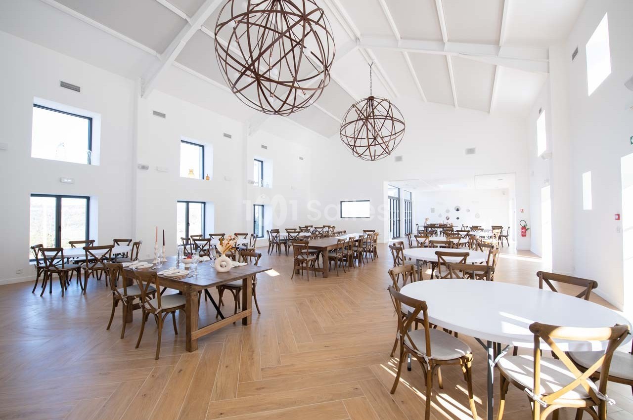 Salle lumineuse avec de grandes fenêtres, des tables en bois et des chaises, décorée de suspensions métalliques.