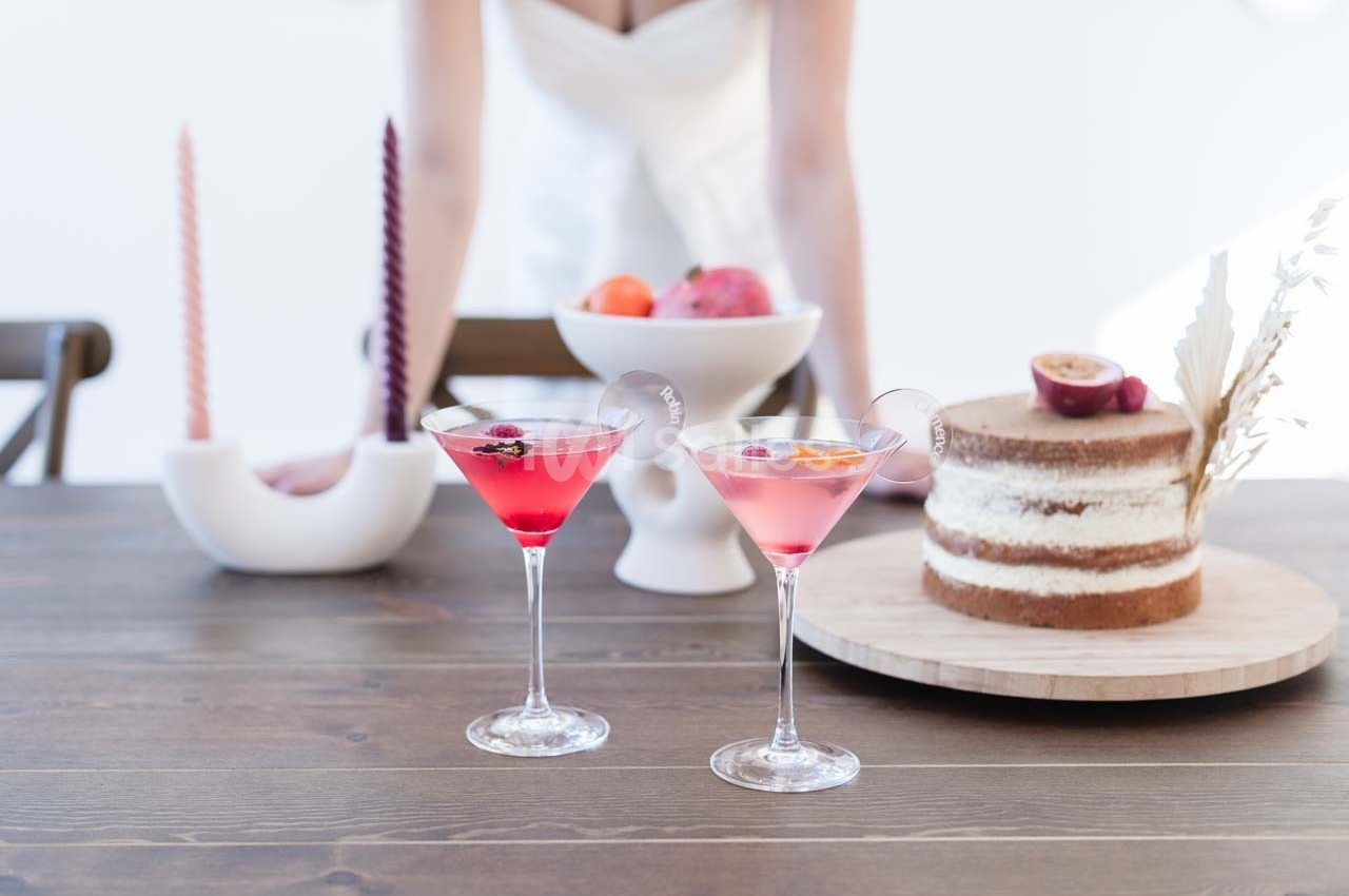 Deux cocktails roses sur une table en bois, accompagnés d'un gâteau, de fruits et de bougies décoratives.